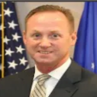 Portrait of a man in a suit with American flags in the background.
