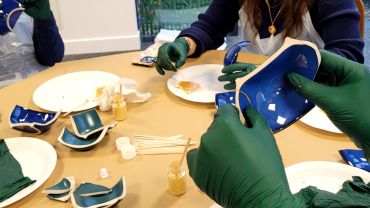 Hands in green gloves repairing broken blue ceramics using gold lacquer.