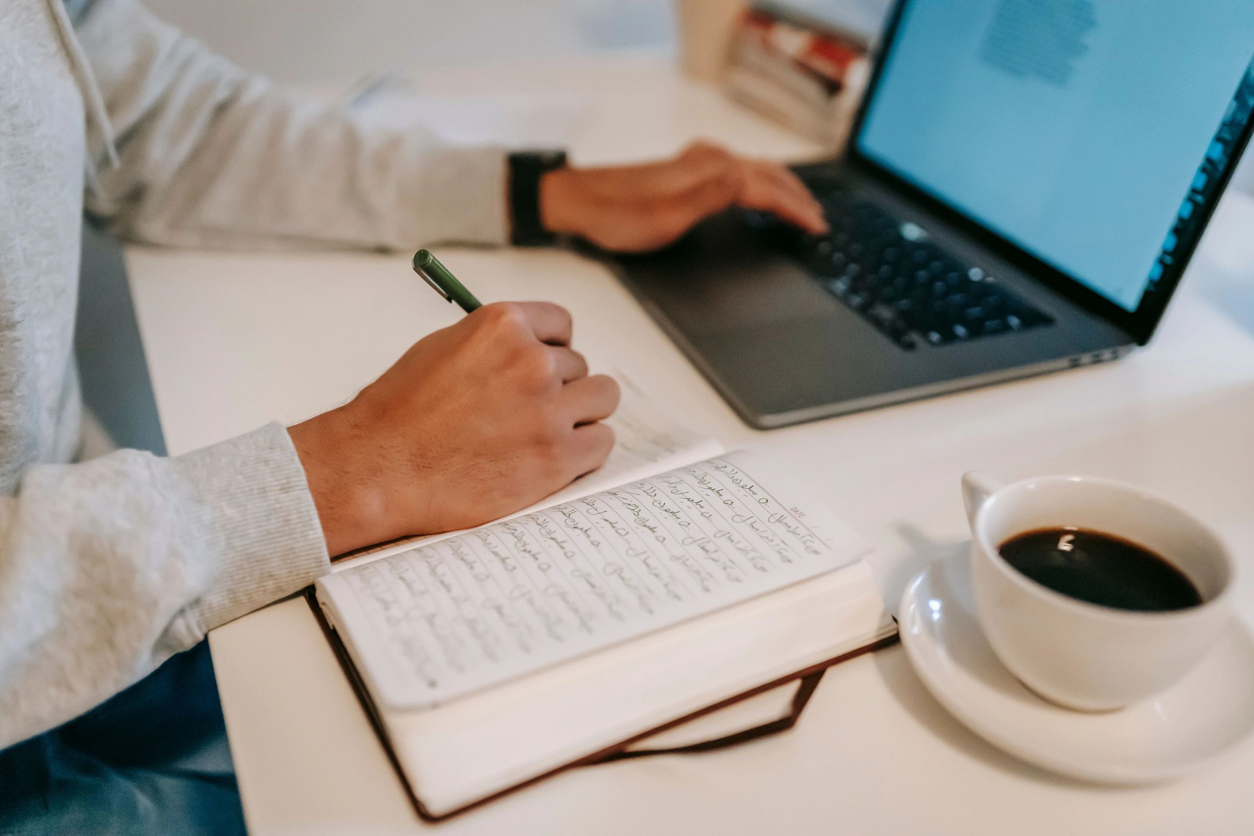 Person writing in notebook while using a laptop with coffee nearby.