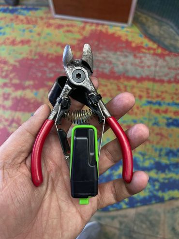 Hand holding a pair of red-handled wire cutters with a black and green attachment.
