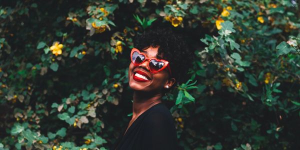 Woman smiling brightly wearing red heart-shaped sunglasses against a leafy backdrop.