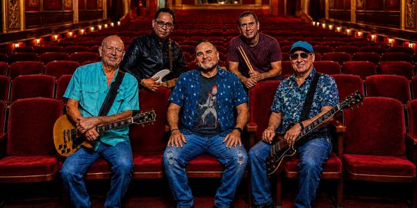 Five musicians posing with guitars in a theater with red seats.