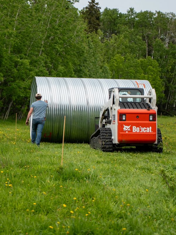 A man guiding a Bobcat skid-steer loader moving a large metal culvert in a grassy field.
