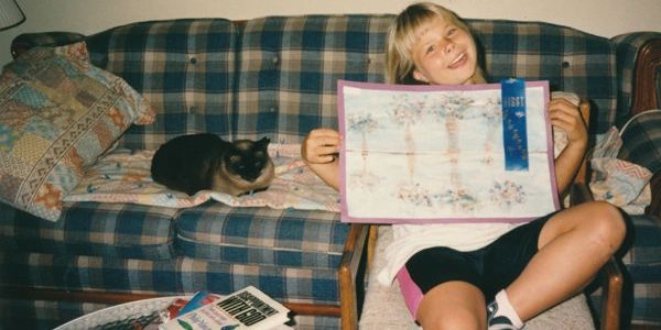 Young girl proudly shows first-place ribbon artwork on a plaid couch beside a resting cat.