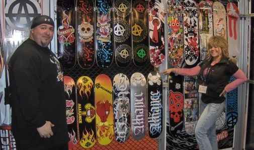 Two people posing by a display of colorful skateboard decks at an Alliance booth.