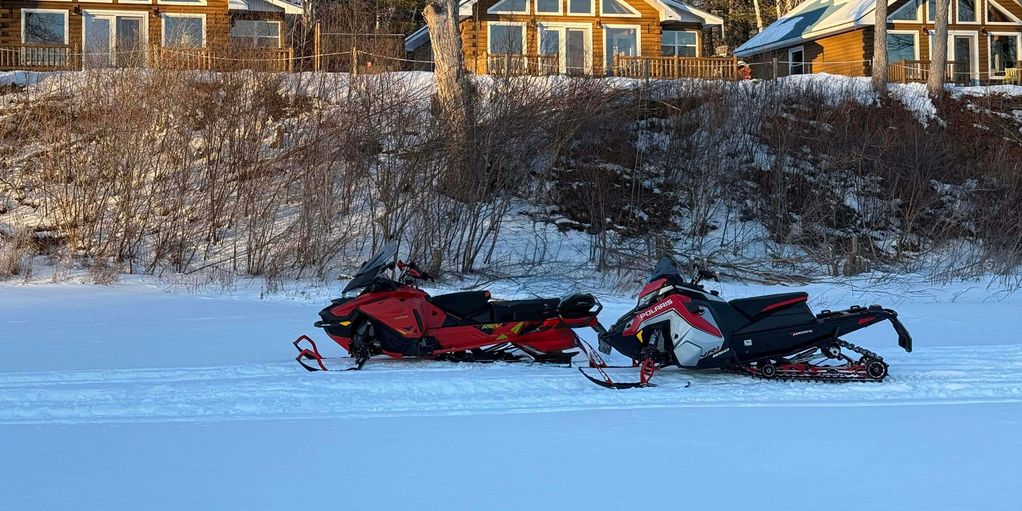 Two snowmobiles parked on snowy ground in front of wooden houses.
