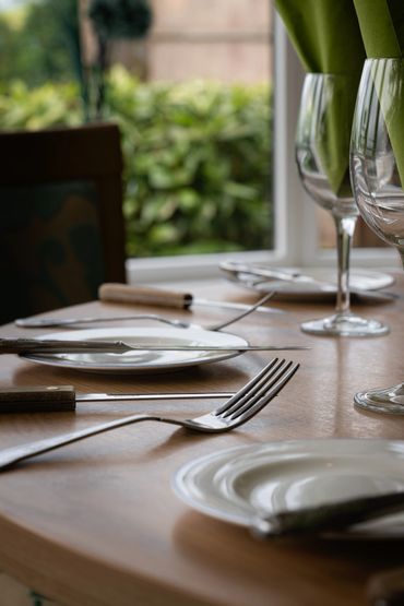 Elegant dining table set with plates, cutlery, and green napkins in wine glasses by a window.