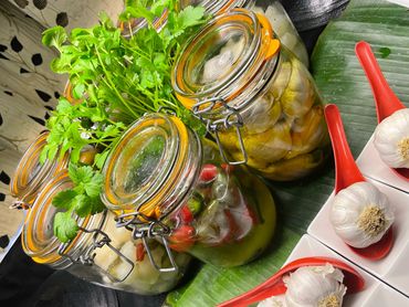 Jars of pickled vegetables and fresh garlic on red spoons arranged on a banana leaf.