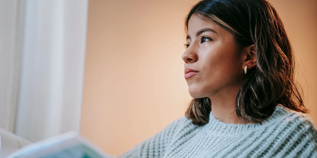 Young woman gazing thoughtfully while holding a book near a window.