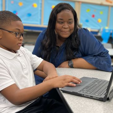 Young boy working on a laptop with a woman observing in a classroom.