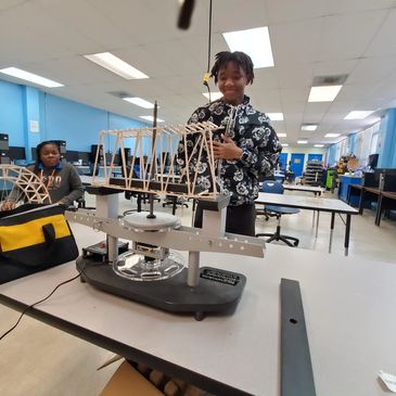 Student presenting a wooden bridge model on a testing machine in a classroom.