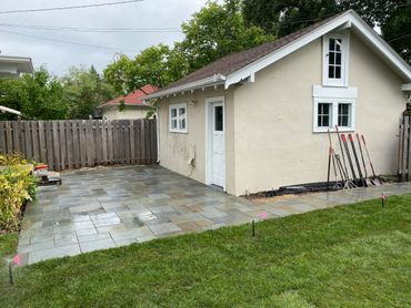 Newly installed wet stone patio beside a small beige building with gardening tools.