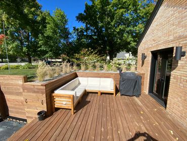 Sunny wooden deck with cushioned seating and a covered grill beside a brick house.