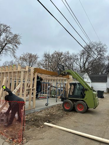 Construction workers build wooden framework using a skid-steer loader on a cloudy day.