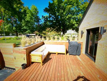Sunny wooden deck with cushioned seating, planter boxes, and a covered grill beside a brick house.