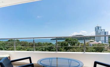 Balcony with chairs and a glass table overlooking trees and the ocean under a clear sky.