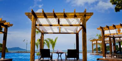Wooden cabanas with lounge chairs by a pool overlooking the ocean under a blue sky.