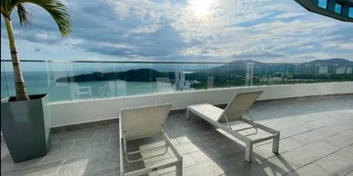 Two lounge chairs on a spacious balcony overlooking the ocean and islands under a bright sun.