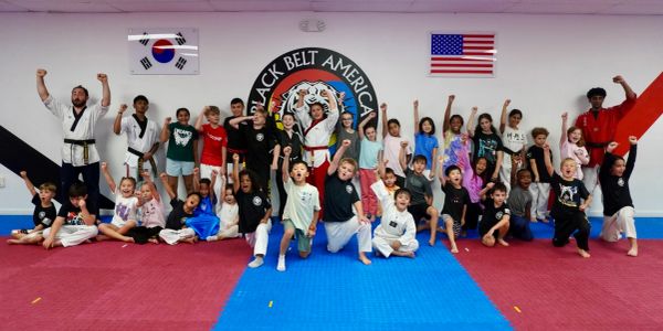 A diverse group of children celebrating in a martial arts studio with flags and logo on the wall.