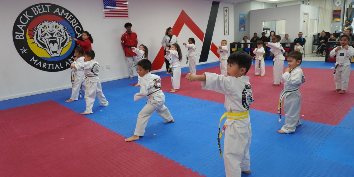 Young children practicing martial arts in a dojo with instructors and spectators.