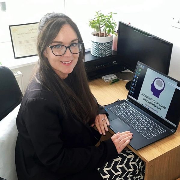 Woman in glasses smiling at laptop showing hypnotherapy website.