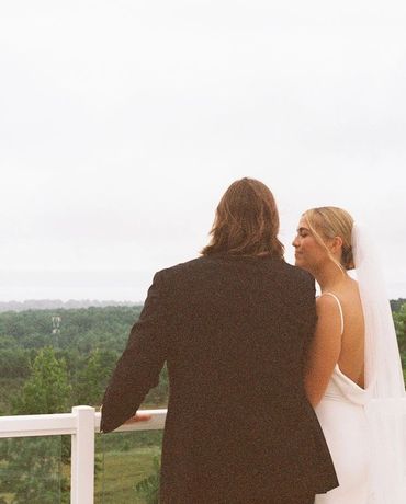 Newlyweds in elegant attire gaze over a scenic landscape from a balcony.
