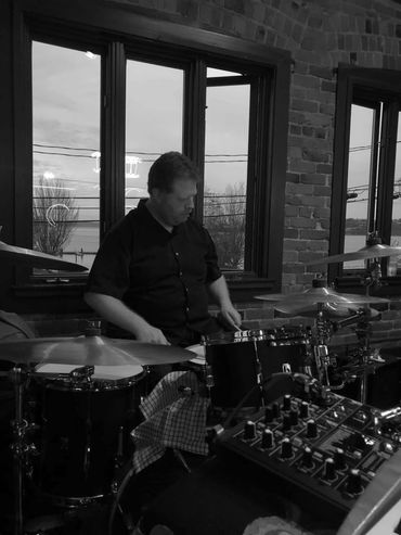 Man playing drums in a cozy room with large windows and brick walls.