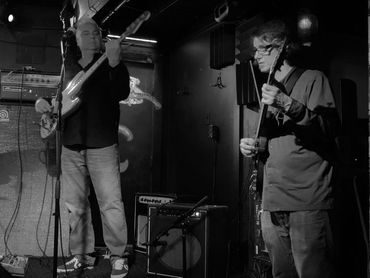 Two musicians play guitars on a dimly lit stage with colorful lighting.