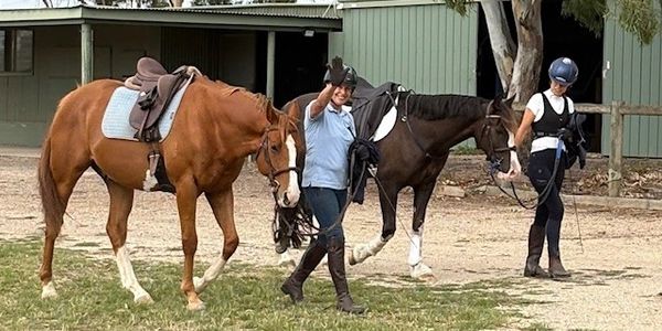 Two people leading horses on a ranch, one person waving.