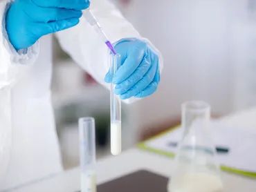 Scientist in blue gloves pipetting a liquid into a test tube in a lab.