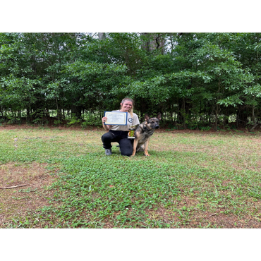 Woman kneels with a German Shepherd holding a certificate and trophy outdoors.