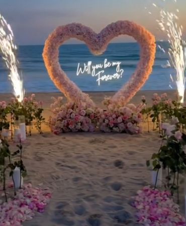 Romantic beach proposal setup with heart-shaped flowers and fireworks.