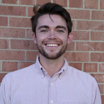 A smiling young man in a light button-up shirt stands against a brick wall.