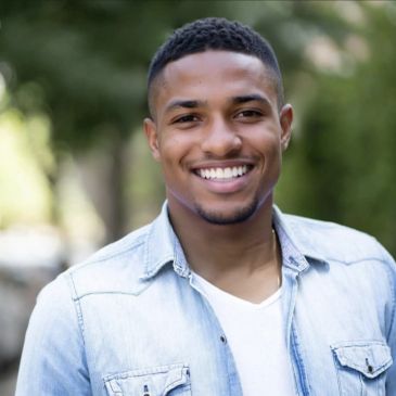 A young man smiling outdoors in casual denim jacket.