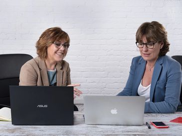 Dos profesionales ejecutivas colaborando eficazmente en un entorno de trabajo, demostrando liderazgo y habilidades de equipo con laptops ASUS y Apple.