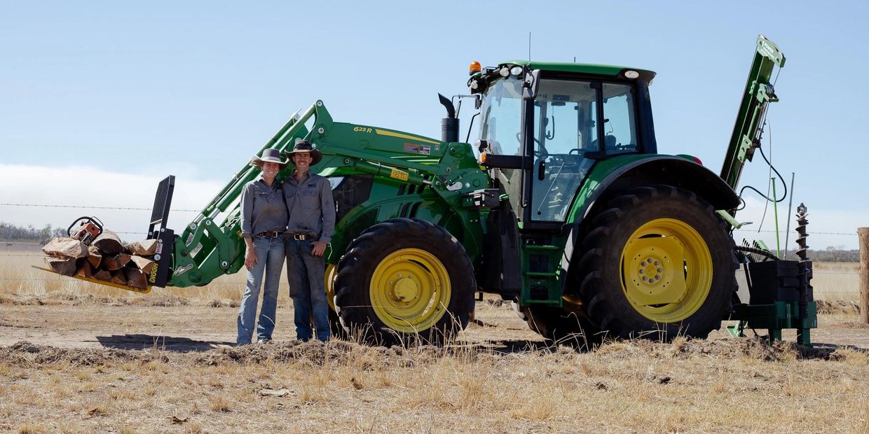 Two farmers standing next to a green tractor with wood logs in a rural field.