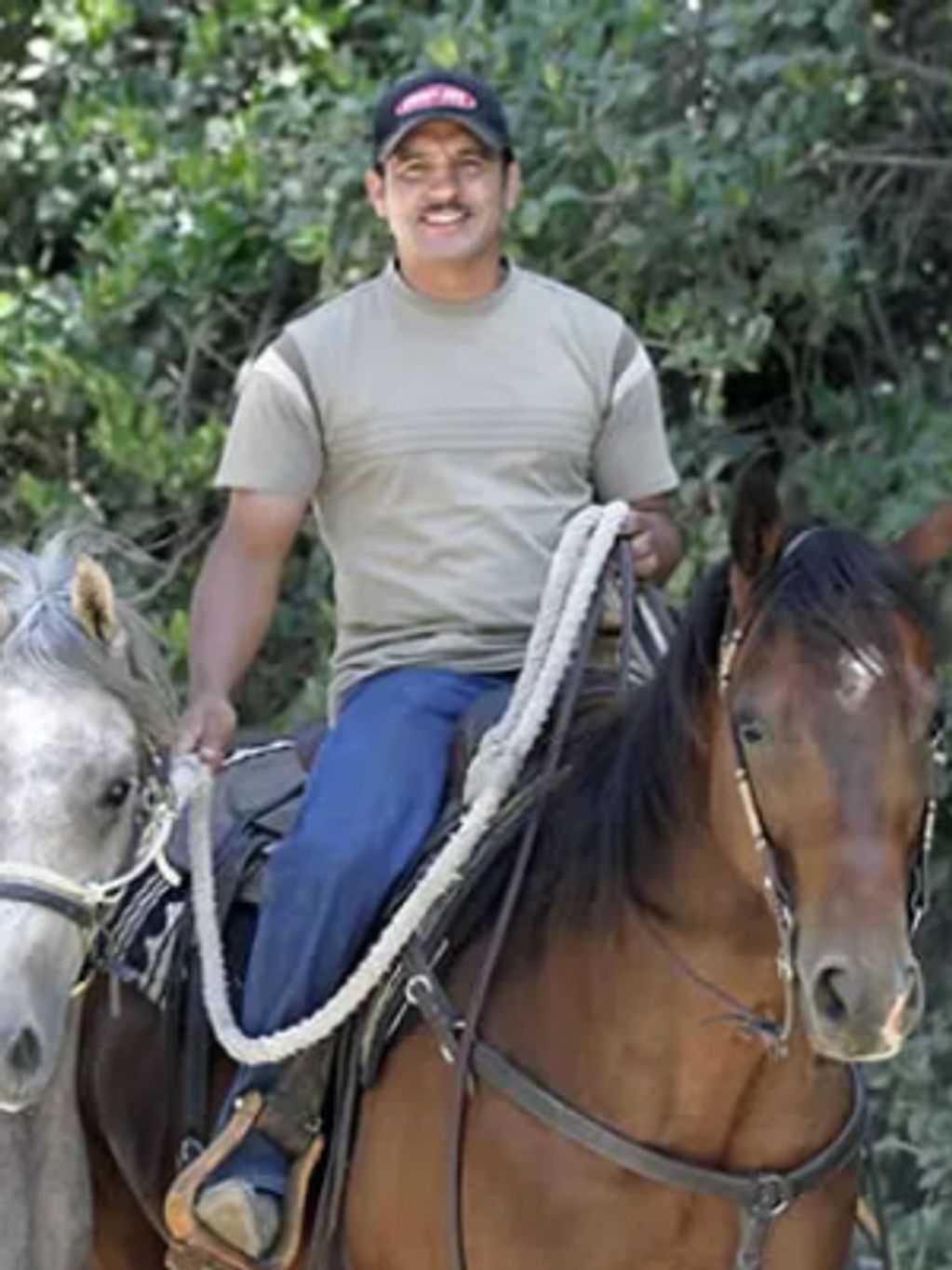 Man smiling while riding a brown horse and holding a white horse's reins.