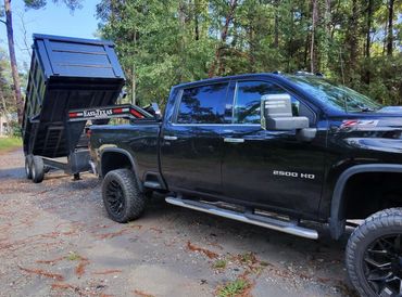 Black 2500 HD truck towing an East Texas dump trailer in a forested area.