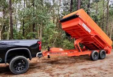 Black pickup truck towing an orange RawMaxx dump trailer with raised bed in a forested area.