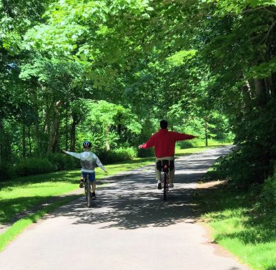 Two people riding bikes on a tree-lined path with arms outstretched.