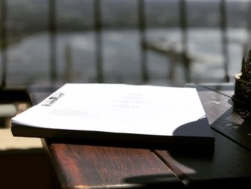 A stack of papers lies on a wooden table by a window with a view of a body of water and a bridge in the distance. Sunlight casts shadows across the table and papers. A decorative metallic item is partially visible on the right side of the table.