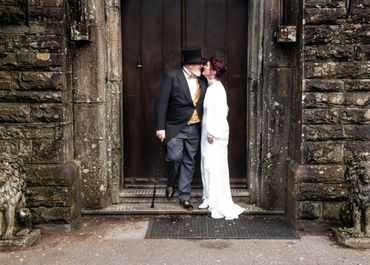 An older couple in vintage attire sharing a kiss in front of a stone building.