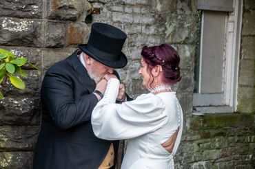 Man in formal wear kisses the hand of a woman in a white dress against a stone wall.