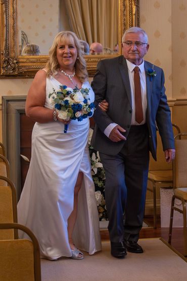 Bride in white dress holding a bouquet walks arm-in-arm with an elderly man in a suit.