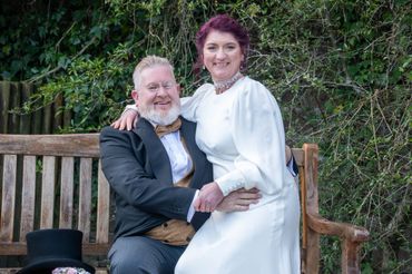 A happy couple dressed in vintage wedding attire sitting on a wooden bench.