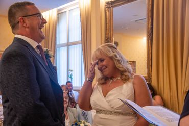 Bride wipes tear while groom laughs during wedding ceremony indoors.
