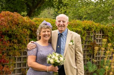 Happy elderly couple posing outdoors, holding a bouquet of white flowers.