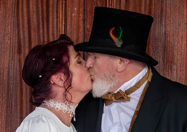 An elderly couple shares a kiss in vintage attire against a wooden backdrop.