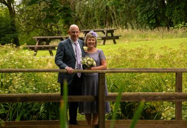Smiling couple in formal attire poses on a wooden bridge in a lush garden.