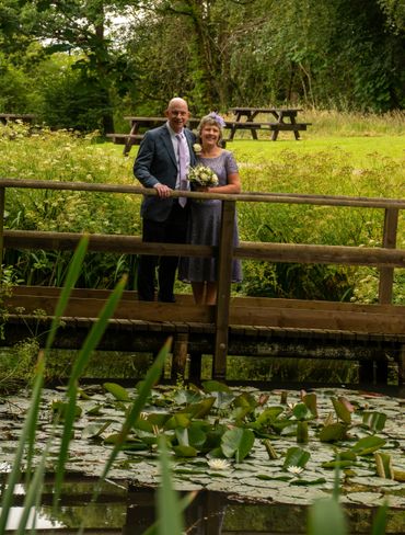 Couple standing on a wooden bridge over a lily pond in a lush garden.